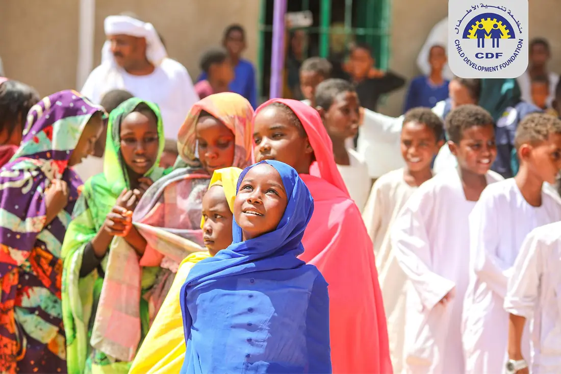 Girls and women in brightly coloured headscarves at a community gathering, with boys in white jalabiyas behind them