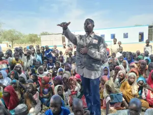 موظف ميداني يتحدث ويشير بيده إلى حشد كبير من الأطفال يجلسون أمامه في العراء