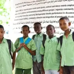 خمسة تلاميذ بقمصان مدرسية خضراء يحملون حقائب الظهر ويقفون أمام لافتة مشروع كبيرة باللغتين
