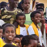 مجموعة من الأولاد المبتسمين في الهواء الطلق، اثنان في المقدمة يرتديان صديريات صفراء فوق جلابيب بيضاء
