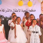 فتيات بفساتين بيضاء يغنين على المسرح ويلوّحن بأيديهن، وخلفهن لافتة المنظمة وبالونات