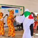فتيات بأثواب برتقالية مزهرة يؤدين رقصة على مسرح مدرسي، وإحدى الراقصات ملتحفة بعلم السودان