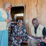 موظفون من المنظمة يحملون استمارات وأقلاماً يجرون مقابلة مع امرأة تجلس على سرير أمام بيت من الطين