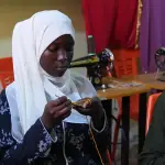 شابة بحجاب أبيض تشتغل بالكروشيه بخيط برتقالي، وخلفها ماكينات خياطة على الطاولات