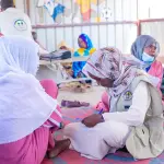 موظفة من المنظمة تجلس على الحصيرة وتدوّن ملاحظات بينما تتحدث النساء حولها