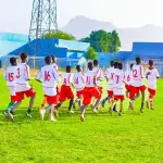 أولاد بزي كرة القدم الأبيض والأحمر يركضون في ملعب أخضر، والجبال خلف الاستاد